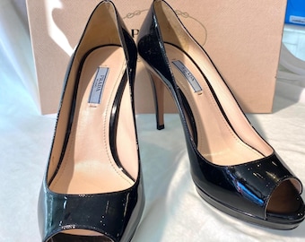 May include: A pair of black patent leather peep-toe high heels. The shoes have a platform sole and a stiletto heel. The Prada logo is visible on the inner sole. The shoes are displayed in front of a Prada box.