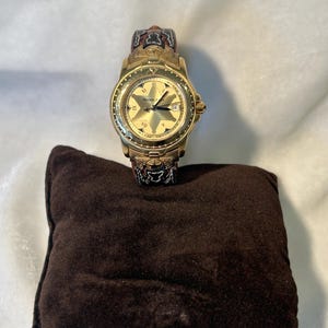 Puede incluir: Un reloj dorado con esfera en forma de estrella, n&uacute;meros romanos y visualizaci&oacute;n de fecha. La correa del reloj tiene un dise&ntilde;o con detalles rojos, negros y dorados. Descansa sobre un peque&ntilde;o coj&iacute;n cuadrado marr&oacute;n.