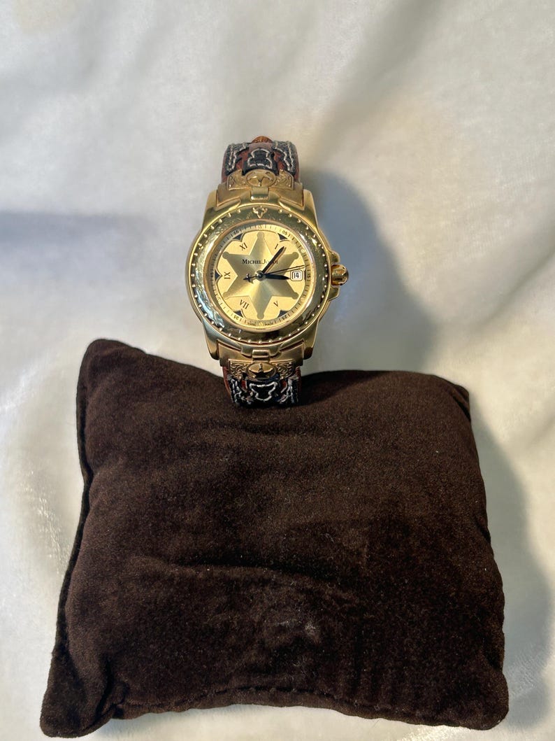 Puede incluir: Un reloj dorado con esfera en forma de estrella, n&uacute;meros romanos y visualizaci&oacute;n de fecha. La correa del reloj tiene un dise&ntilde;o con detalles rojos, negros y dorados. Descansa sobre un peque&ntilde;o coj&iacute;n cuadrado marr&oacute;n.
