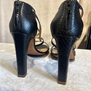 May include: Pair of black high-heeled sandals with a snakeskin texture. The heels are thick and square, with a zipper closure at the back. The straps are thin and delicate, and the soles are a light brown color.