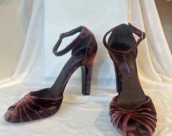 May include: A pair of dark brown velvet high-heeled sandals featuring a peep-toe design with gathered fabric detailing at the front, an adjustable ankle strap with a gold-tone buckle, and a high block heel. The interior insole is labeled Ralph Lauren.