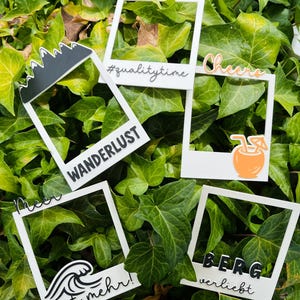 Könnte beinhalten: Fünf weiße rechteckige Fotorahmen mit verschiedenen Verzierungen. Einer hat eine schwarze Bergspitze und den Text WANDERLUST. Ein anderer hat #qualitytime. Einer zeigt das Wort Cheers und ein orangefarbenes Getränk. Einer hat eine Welle und den Text meer ist mehr!. Einer hat den Text BERG verliebt.