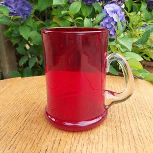 May include: A ruby red glass mug with a clear handle. The mug has a rounded base and a straight, cylindrical body. The background features green foliage and purple flowers, suggesting an outdoor setting. The mug is likely used for beverages.