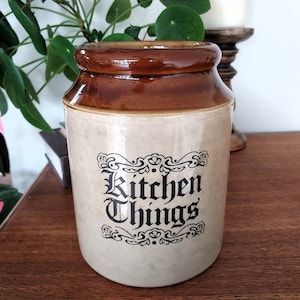 May include: A vintage-style kitchen storage jar with a brown glazed top and a cream-colored base. The jar features the words "Kitchen Things" in a decorative black font. The jar is made of ceramic and is likely used for storing dry goods.