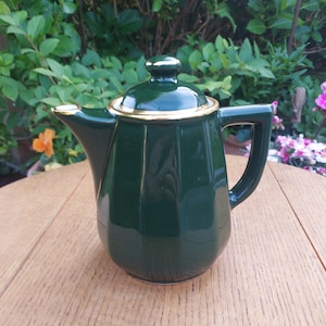 May include: A dark green ceramic teapot with a gold rim and handle. The teapot has a lid with a small knob and is sitting on a wooden table. The background features green foliage and flowers.