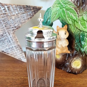 May include: A clear glass jar with a silver lid and a small spout. The jar has a faceted design and is sitting on a wooden surface.