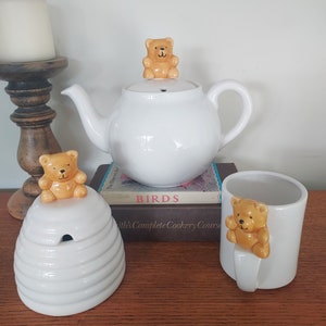May include: White ceramic teapot, honey pot, and mug with brown bear figurines. The teapot has a bear figurine on the lid. The honey pot is shaped like a beehive. The mug has a bear figurine on the handle. The items are on a wooden surface.
