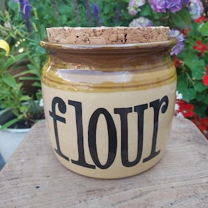 May include: A brown ceramic canister with a cork lid and the word "flour" printed in black letters. The canister is sitting on a wooden surface.