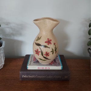 Peut inclure: Un vase en céramique beige avec une finition mouchetée et une large ouverture. Le vase est décoré de fleurs rouges peintes à la main et de feuilles vertes. Le vase est posé sur deux livres, l'un intitulé "Birds" et l'autre intitulé "Delia Smith's Complete Cookery Course."