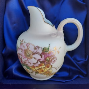 Könnte beinhalten: Ein kleiner, weißer Porzellan-Krug mit Blumenmuster. Der Krug hat einen goldenen Rand und einen Henkel. Er steht auf einem blauen Samttuch.