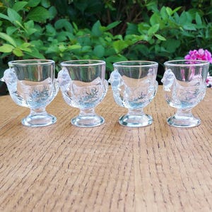 May include: Four clear glass egg cups with a chicken design. The egg cups are on a wooden surface with green foliage in the background. Each cup has a round base and a detailed chicken figure on the side.