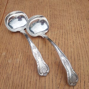 Op de afbeelding: Twee zilveren serveerscheppen met sierlijke handvatten. De scheppen hebben een gepolijste afwerking en zijn in uitstekende staat.