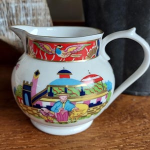May include: White ceramic creamer with a decorative design. The creamer features a red band with a bird and gold accents. The body of the creamer has a colorful scene with figures and buildings. The handle is white and curved.