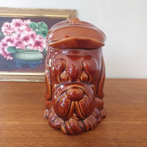 May include: A brown ceramic dog-shaped cookie jar with a lid shaped like a cap. The jar has a glossy finish and detailed features, including a prominent snout and floppy ears. The jar is sitting on a wooden surface.
