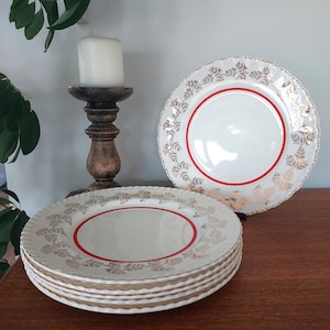 May include: A stack of six white ceramic plates with a gold and red floral design. The plates have a scalloped edge and a red band around the center.