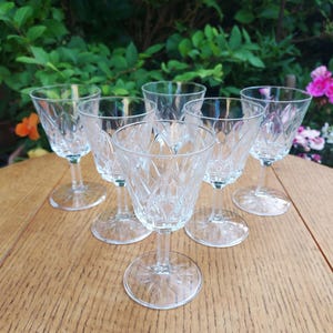 May include: A set of six clear crystal wine glasses, each with a decorative diamond and leaf design. The glasses stand on stemmed bases. They are arranged on a wooden table, with a backdrop of green leaves and pink flowers.