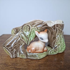 Peut inclure: Un pot de fleurs en céramique en forme de tronc d'arbre avec une figurine de faon à l'intérieur. Le pot est de couleur marron, vert et blanc, avec des textures détaillées de feuilles et d'écorce. Le faon est brun clair avec des taches blanches.