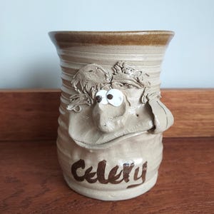 Op de afbeelding: Een beige keramische keukengerei houder met een grillig gezichtsontwerp. De houder heeft een brede rand en het woord "Celery" is in bruin schrift op de voorkant geschreven. Het gezicht heeft grote witte ogen en een neus.