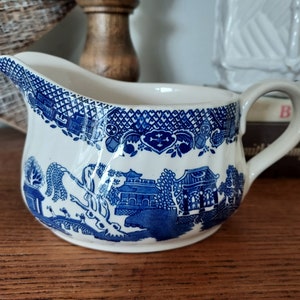 May include: A white ceramic creamer with a blue and white willow pattern. The creamer has a handle and a spout.