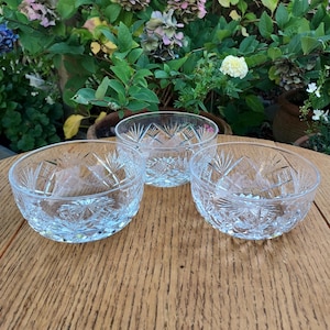 May include: Three clear glass bowls with intricate starburst and diamond patterns. The bowls are arranged on a wooden table, with a backdrop of green foliage and flowers. The bowls are likely for serving desserts or snacks.