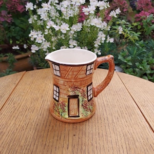 May include: A decorative ceramic jug featuring a house design. The jug is brown with a white interior and a handle. The house design includes windows, a door, and brick detailing. The jug is on a wooden table.