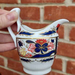 May include: A small white porcelain creamer with a blue, red, and gold floral pattern. The creamer has a handle and a spout.