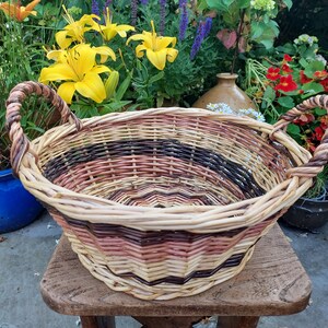 Peut inclure: Un panier en osier tressé avec deux anses. Le panier est de couleur marron, rose et naturel et présente un motif rayé. Le panier est posé sur un tabouret en bois.