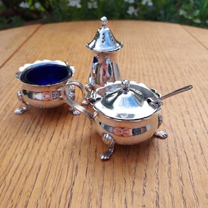 May include: A trio of antique silver serving dishes. The set comprises a small bowl with a cobalt blue interior, a lidded sugar bowl with a spoon, and a salt shaker. Each piece features ornate detailing and rests on small, decorative feet.