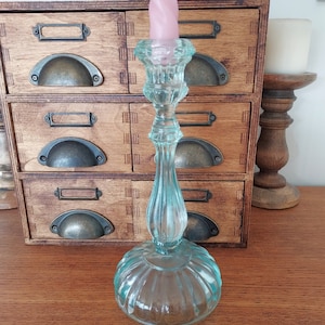 May include: A light blue glass candle holder with a fluted base and a twisted pink candle. The candle holder is sitting on a wooden surface.
