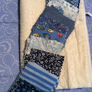 May include: A stack of square fabric swatches in various shades of blue, featuring patterns such as stripes, florals, paisley, and small teacup motifs, layered over a rectangular piece of thick, cream-colored, textured felted fabric.