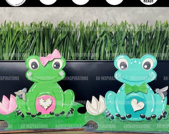 Peut inclure: Deux figurines de grenouilles, l'une verte et l'autre bleue, assises sur un lit de feuilles vertes. Les deux grenouilles ont un cœur sur le ventre et tiennent une fleur. La grenouille verte a un nœud rose sur la tête. La grenouille bleue a un nœud papillon vert.