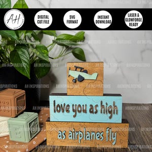 Puede incluir: Un conjunto de bloques de madera de tres niveles con el texto "love you as high as airplanes fly". El bloque superior presenta un pequeño avión con nubes. Los bloques están pintados en tonos de marrón, azul y verde.