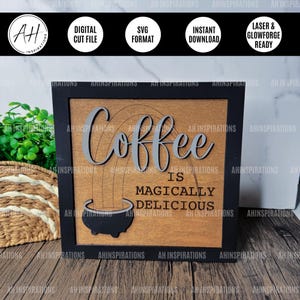 Pode incluir: Uma placa de madeira com moldura preta e o texto "Coffee is magically delicious" em fonte cinza. Um desenho de arco-íris está atrás do texto e uma silhueta preta de uma cafeteira está abaixo do texto.