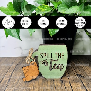 Puede incluir: Un letrero de madera con forma de taza de té con el texto "Spill the Tea" en un esquema de colores verde y marrón. Un pequeño amuleto de tetera de madera cuelga del letrero.