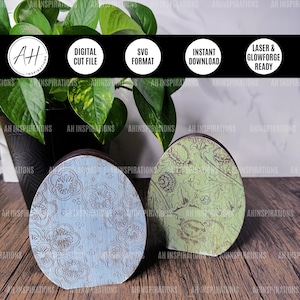 May include: Two wooden Easter eggs, one painted light blue with a white floral pattern and the other painted green with a brown floral pattern. The eggs are sitting on a wooden surface.