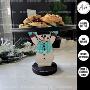 May include: A wooden snowman cake stand with a black base and a teal and white snowman design. The snowman is holding a black tray with cookies on it. The text "DIGITAL CUT FILE" is visible in the image.