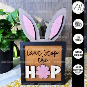 Può includere: Un cartello di legno con un design a forma di orecchie di coniglio e il testo "Can't Stop the Hop" in lettere bianche e nere. Il cartello ha un fiore rosa al centro della parola "Hop".