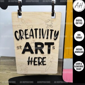 May include: A wooden sign with the words "Creativity Starts Here" in black lettering. The sign is decorated with paint splatters and a paintbrush. The sign is hanging from a string.