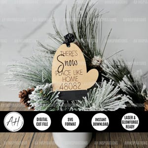 May include: A wooden Christmas ornament shaped like a mitten with the text "There's Snow Place Like Home 48082" engraved on it. The ornament is brown with black trim and is sitting on a bed of pine branches.