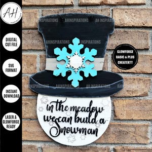 Può includere: Un portapacchi per porta in legno bianco a forma di pupazzo di neve con un cappello a cilindro nero, un fiocco di neve blu e il testo "in the meadow we can build a snowman".