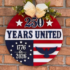 May include: A round wooden sign with a red, white, and blue design. The sign features the text "250 YEARS UNITED" and "1776 to 2026". A blue eagle and white stars are also included. The sign is decorated with a cream-colored bow and greenery.