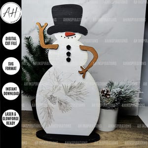 May include: A white wooden snowman with a black top hat, brown arms, and a black and white pine bough design. The snowman is standing on a black base.