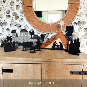 May include: A wooden nativity scene with black silhouettes of figures, buildings, and animals. The scene includes a star, an angel, and the text "Oh Holy Night". The figures are arranged on a wooden surface in front of a decorative mirror.