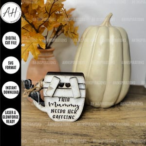 Könnte beinhalten: Ein Holzschild in Form einer Kaffeetasse mit dem Text "This Mummy Needs Her Caffeine" darauf. Das Schild steht auf einer Holzoberfläche neben einem weißen Kürbis und einer Vase mit Herbstblättern.