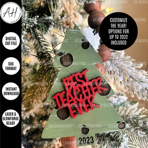 May include: A green Christmas tree ornament with a red "Best Teacher Ever" sign and the year 2023. The ornament is decorated with red apples and has a rustic woodgrain texture.