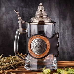 May include: A clear glass beer stein with a silver lid and handle. It has a leather wrap with a metal emblem that reads "JACK GROOMSMAN". The stein is on a wooden surface with hops and barley.