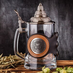 May include: A clear glass beer stein with a silver lid and handle. The stein has a brown leather wrap with a metal emblem that reads "THIS DAD DESERVES BEER". The stein is on a wooden surface.