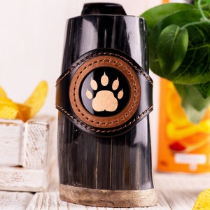 Può includere: Una tazza in corno nero con una fascia in pelle marrone e un design a forma di impronta di zampa. La tazza è appoggiata su una superficie bianca.