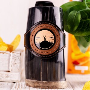 Può includere: Una boccale da bere in corno nero con una fascia in pelle marrone e un'intarsio circolare che presenta la silhouette di un cervo in piedi su una collina. L'intarsio è circondato da una fascia in pelle marrone con cuciture.