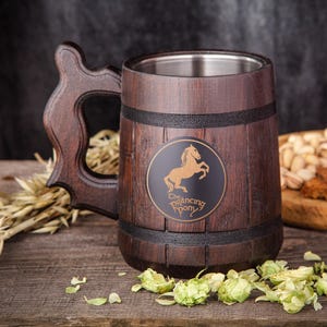 May include: A wooden beer mug with a stainless steel interior. The mug has a dark brown finish with a decorative handle and a circular emblem featuring a horse. The words "The Prancing Pony" are below the horse. The mug is on a wooden surface.